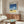 May include: Framed poster featuring blue sky with white clouds over yellow sand and vintage dark car, displayed above wooden sideboard in modern living room with white curved chair, glass coffee table, and warm lighting creating shadows on neutral walls.