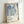 Framed exhibition poster featuring abstract blue and white flowing organic shapes with dotted patterns and circular motifs. The artwork shows fluid, elongated forms in various shades of blue against a cream background. Below the artwork is text reading "YAYOI KUSAMA" with Japanese characters and "Tokyo, Japan 1984". The poster is displayed in a light wooden frame leaning against a white wall, casting a soft shadow.