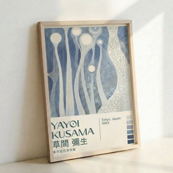Framed exhibition poster featuring abstract blue and white flowing organic shapes with dotted patterns and circular motifs. The artwork shows fluid, elongated forms in various shades of blue against a cream background. Below the artwork is text reading "YAYOI KUSAMA" with Japanese characters and "Tokyo, Japan 1984". The poster is displayed in a light wooden frame leaning against a white wall, casting a soft shadow.