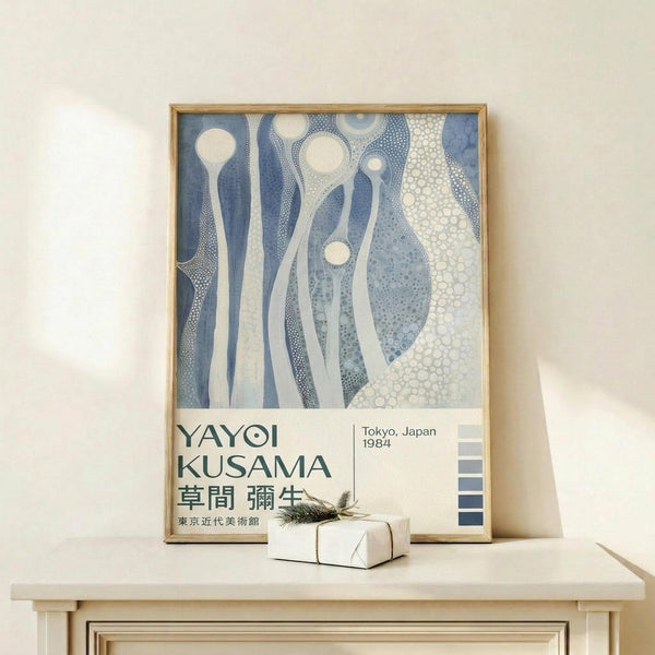 May include: Framed exhibition poster featuring abstract blue and white organic forms with dotted patterns, displayed on light wood surface next to small wrapped gift with twine. Poster shows flowing, cellular-like shapes in various shades of blue against cream background, with Japanese text at bottom and color palette strip on right side.