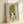 May include: Framed art print leaning against white wall with natural light shadows. Features anime-style character with oversized golden blonde beehive hair, large dark eyes, and yellow-green outfit on soft blue background. Light wood frame, approximately 8x10 size, positioned at slight angle showing dimensional depth.