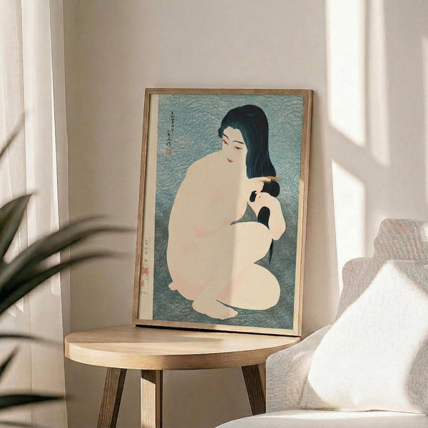 May include: Japanese woodblock print artwork displayed in thin wooden frame on round wooden side table next to beige upholstered chair. Print shows woman with long black hair combing hair of smaller figure against blue-green textured background. Soft natural lighting creates shadows on white wall. Green plant leaves visible in foreground.
