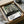 May include: Framed art print in cardboard shipping box with protective tissue paper and wooden ruler props. Abstract poster features vertical black drips over cream and sage green organic shapes with dotted patterns at bottom. White label shows artist name and Tokyo, Japan 1984. Minimalist contemporary art style with earth tone color palette.