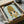 May include: Framed artwork being unpacked from cardboard shipping box with protective wrapping. The print shows a cute cartoon-style character with large green eyes, blonde hair in a beehive style, and yellow clothing against a pale green background. The piece sits in a black frame surrounded by white tissue paper and wooden frame corners for protection.