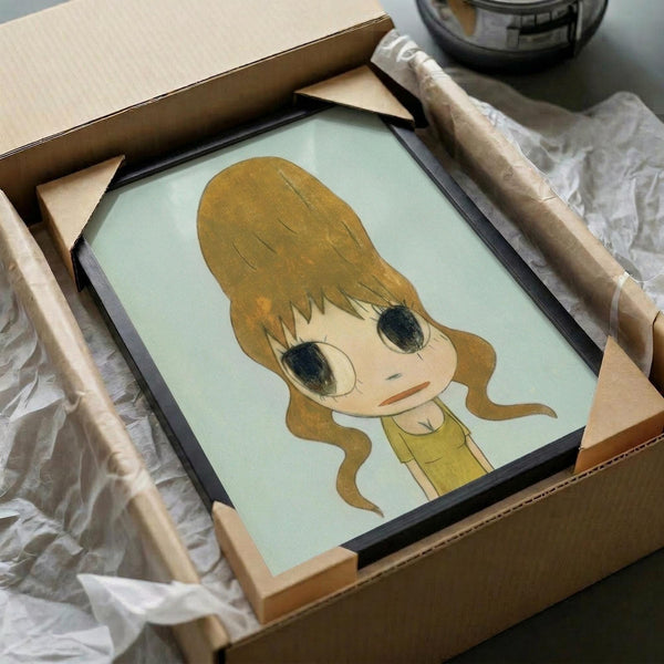 May include: Framed artwork being unpacked from cardboard shipping box with protective wrapping. The print shows a cute cartoon-style character with large green eyes, blonde hair in a beehive style, and yellow clothing against a pale green background. The piece sits in a black frame surrounded by white tissue paper and wooden frame corners for protection.