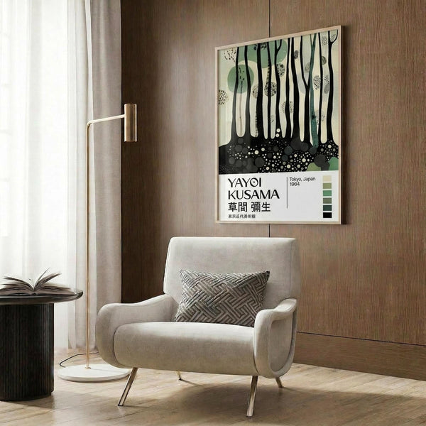 May include: Modern art poster featuring abstract forest scene with black tree trunks, green circles, and dotted patterns on cream background, displayed in light wood frame on warm wood-paneled wall. Beige upholstered armchair with geometric patterned pillow sits below, alongside brass floor lamp and dark round side table with open book.