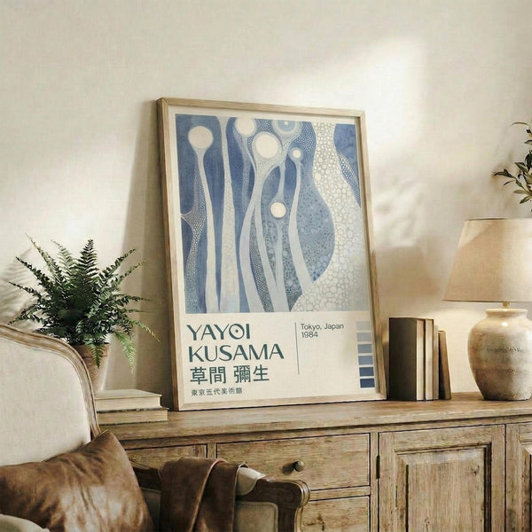 May include: Framed poster with abstract blue and white organic shapes displayed on rustic wooden sideboard alongside fern plant, ceramic vase, and books in warm-toned living room setting with beige armchair visible