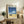 May include: Framed poster with blue sky, white clouds, ocean waves, and dark car on sandy beach, displayed on rustic wooden sideboard with beige chair, potted fern, ceramic vase, and stacked books in neutral-toned living room setting