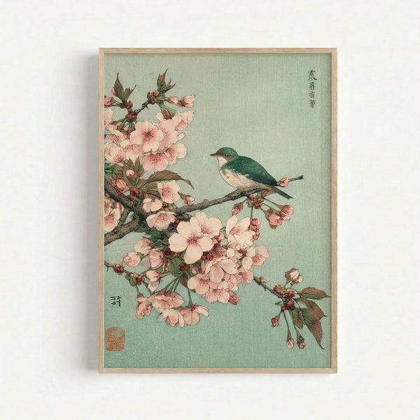 Framed Japanese woodblock print featuring a small green and white bird perched on a flowering cherry blossom branch with delicate pink petals against a soft mint green background. The artwork includes traditional Japanese calligraphy and a red artist's seal in the corners, displayed in a light wooden frame against a white wall.