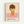 Framed poster featuring a minimalist illustration of a young child with brown bob haircut and closed eyes, wearing an orange dress with white daisy, standing against a pink and cream background. The artwork has a sweet, melancholic style with soft colors. Below the image is text reading "YOSHITOMO NARA" in black letters with Japanese characters and exhibition dates from 1992. The poster is displayed in a light wood frame against a white wall.