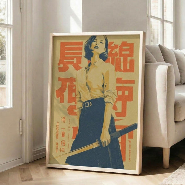 May include: Framed vintage-style poster featuring a woman with short dark hair wearing a yellow blouse and blue skirt, holding a katana sword. The artwork has a retro Japanese aesthetic with bold orange text characters on a cream background. White wooden frame is propped against a beige sofa in a bright living room with white walls and windows.