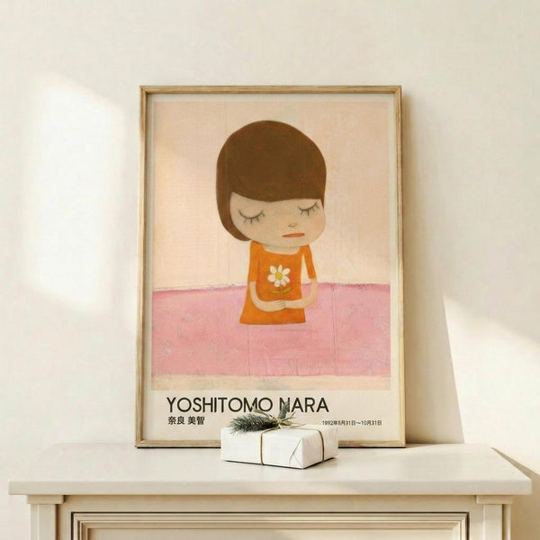 May include: Framed poster displaying a minimalist illustration of a young child with brown bob haircut wearing an orange dress with white daisy, eyes closed, standing on pink ground against cream background. Text reads "Yoshitomo Nara" with Japanese characters and dates. Light wood frame sits on white wooden surface next to small wrapped gift with evergreen sprig against white wall.
