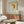 May include: Framed poster featuring child-like figure with orange cap emerging from green rippled water against pale background, displayed above mid-century walnut credenza in modern living room with white curved chair, glass coffee table, and neutral walls with natural lighting