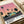 Retro-style illustration poster featuring tall palm trees silhouetted against a pink and orange sunset sky. A mid-century modern turquoise house sits beside a dark road with a vintage car. Bold text reads "HIROSHI NAGAI" at bottom with "TOKYO 1973" and colorful dots in corner. The artwork has a nostalgic 1970s aesthetic with vibrant tropical colors. Poster shown flat on white surface next to rolled paper tube.