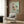 May include: Japanese woodblock print artwork in light wood frame showing pink cherry blossoms and teal bird on mint green background, displayed on warm wood wall paneling above cream upholstered armchair with geometric pillow, brass floor lamp and dark round side table with open book nearby