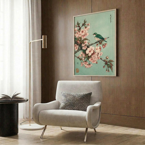 May include: Japanese woodblock print artwork in light wood frame showing pink cherry blossoms and teal bird on mint green background, displayed on warm wood wall paneling above cream upholstered armchair with geometric pillow, brass floor lamp and dark round side table with open book nearby