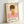 May include: Framed art print leaning against white wall showing minimalist illustration of young girl with brown bob haircut and closed eyes, wearing orange dress with white daisy, sitting on pink surface. Light wooden frame surrounds cream-colored poster with black text at bottom including artist name and Japanese characters.