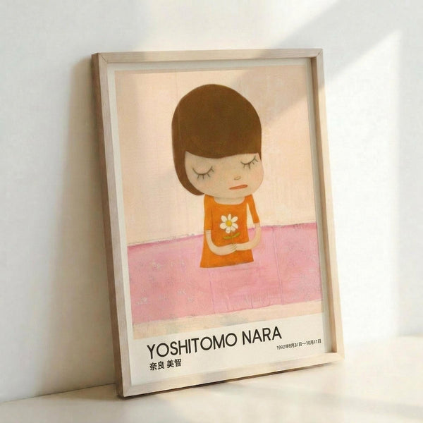May include: Framed art print leaning against white wall showing minimalist illustration of young girl with brown bob haircut and closed eyes, wearing orange dress with white daisy, sitting on pink surface. Light wooden frame surrounds cream-colored poster with black text at bottom including artist name and Japanese characters.