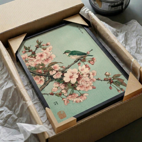 May include: Framed Japanese woodblock print featuring pink cherry blossoms on branches with a small green and white bird perched among the flowers, set against a soft mint green background. The artwork shows delicate botanical details and includes traditional Japanese characters and a red seal stamp. The print is displayed in a dark frame, partially unpacked from a cardboard shipping box with protective wrapping materials visible around it.