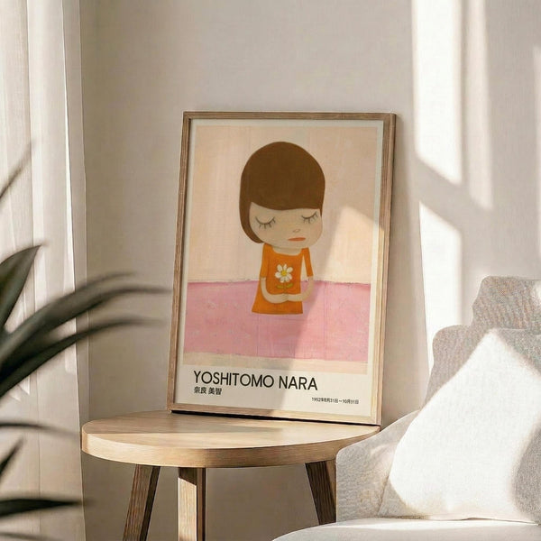 May include: Framed poster featuring a stylized illustration of a young girl with brown bob haircut and closed eyes, wearing an orange dress with white daisy, sitting on pink checkered floor against cream background. Text reads "YOSHITOMO NARA" with Japanese characters and exhibition dates. Frame sits on round wooden side table next to beige upholstered chair with white pillow, green plant leaves visible, natural sunlight casting shadows on white wall.