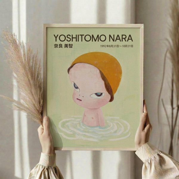 May include: Hands holding a framed poster featuring a minimalist illustration of a child-like figure with orange hair emerging from concentric ripples in water against a pale green background. The artwork has Japanese text and exhibition dates from 1992. Dried pampas grass visible on the left side of the neutral-toned room setting.