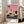 May include: Framed poster leaning against white wall next to beige sofa in bright living room with large windows. Artwork shows turquoise building with palm trees against pink-orange gradient sunset sky, dark road in foreground. Text reads "HIROSHI NAGAI" and "TOKYO 1973" with colored dots. Light wood frame, approximately 2-3 feet tall.