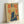 May include: Framed vintage-style poster featuring a woman in samurai attire holding a katana sword, rendered in yellow, blue and red color palette with Japanese text, displayed in light wood frame leaning against white wall corner