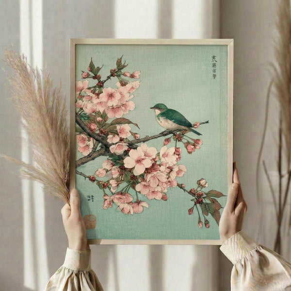 May include: Hands holding a framed Japanese woodblock print featuring pink cherry blossoms and a small green and white bird perched on branches against a soft mint green background. The artwork displays traditional Asian calligraphy in the upper right corner and appears to have a red artist's seal. The print is shown in a thin light wood frame, with dried wheat stalks visible to the left and soft natural lighting from nearby windows creating a serene setting.