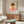 May include: A framed poster featuring a minimalist illustration of a young girl with brown bob haircut and closed eyes, wearing an orange dress with white flower, standing on a pink surface against a cream background. The artwork is displayed above a mid-century modern walnut credenza in a bright living room with white curved chair, oval glass coffee table, and contemporary lighting fixtures.