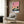 May include: A framed poster showing a mid-century modern house with palm trees against a pink and orange sunset sky, displayed on a wood-paneled wall above a cream upholstered armchair with geometric pillow. The artwork features silhouetted palm trees and a turquoise building with warm evening lighting. A brass floor lamp and dark round side table with an open book complete the modern interior setting.
