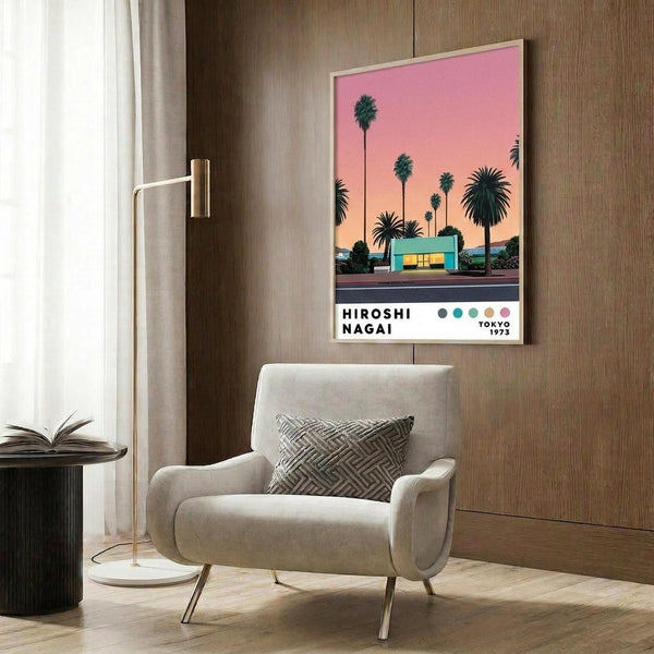 May include: A framed poster showing a mid-century modern house with palm trees against a pink and orange sunset sky, displayed on a wood-paneled wall above a cream upholstered armchair with geometric pillow. The artwork features silhouetted palm trees and a turquoise building with warm evening lighting. A brass floor lamp and dark round side table with an open book complete the modern interior setting.