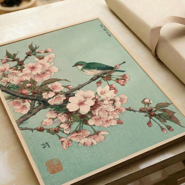 A vintage Japanese woodblock print featuring a small green and white bird perched on a flowering cherry blossom branch. The artwork displays delicate pink sakura flowers with detailed petals and stamens against a soft mint green background. Traditional Japanese characters appear in the upper right corner, with an artist's seal stamp in the lower left. The print is shown in an open art book or portfolio with cream-colored pages, photographed on a white surface with neutral lighting that highlight
