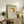 May include: Framed poster featuring a cartoon-style child's head with orange cap emerging from rippling water against pale green background, displayed on rustic wooden sideboard alongside fern plant, ceramic vase, and books in neutral-toned living room setting
