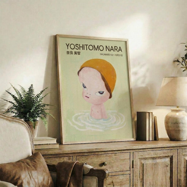 May include: Framed poster featuring a cartoon-style child's head with orange cap emerging from rippling water against pale green background, displayed on rustic wooden sideboard alongside fern plant, ceramic vase, and books in neutral-toned living room setting