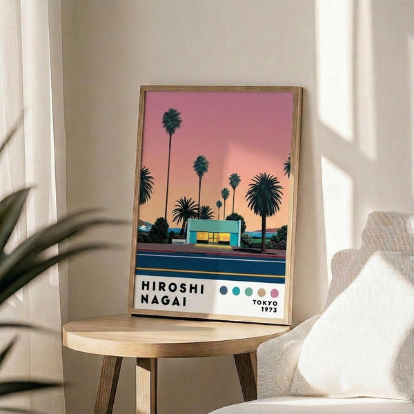 May include: Framed poster displaying palm trees silhouetted against pink and orange sunset sky with small blue building below and road in foreground. Text reads "HIROSHI NAGAI" and "TOKYO 1973" with colored dots. Thin wooden frame sits on round wooden side table next to beige upholstered chair. Soft natural lighting creates shadows on white wall. Plant leaves visible in corner.