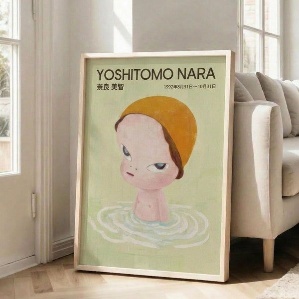 May include: Framed art print featuring a stylized child-like figure with orange hair emerging from rippling water against a soft green background, displayed in light wood frame leaning against white wall next to beige upholstered chair in bright, naturally-lit room with hardwood floors.