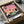 May include: Framed art print featuring palm trees silhouetted against pink and orange sunset sky, with modern turquoise building and road in foreground. Print shows "HIROSHI NAGAI" and "TOKYO 1973" text at bottom with colorful dots. Frame sits in open cardboard shipping box with protective tissue paper wrapping on wooden surface.