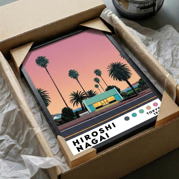 May include: Framed art print featuring palm trees silhouetted against pink and orange sunset sky, with modern turquoise building and road in foreground. Print shows "HIROSHI NAGAI" and "TOKYO 1973" text at bottom with colorful dots. Frame sits in open cardboard shipping box with protective tissue paper wrapping on wooden surface.