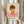 May include: Person holding a framed art print featuring a minimalist illustration of a child with brown bob haircut, closed eyes, wearing an orange dress with white daisy, standing on pink ground against cream background. Light wooden frame, neutral room setting with dried pampas grass visible.