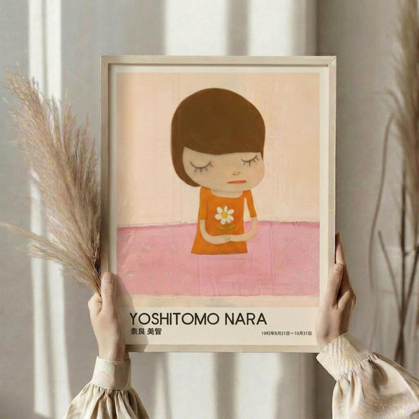 May include: Person holding a framed art print featuring a minimalist illustration of a child with brown bob haircut, closed eyes, wearing an orange dress with white daisy, standing on pink ground against cream background. Light wooden frame, neutral room setting with dried pampas grass visible.