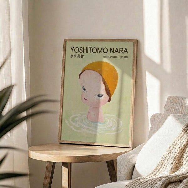 May include: Framed poster featuring a stylized illustration of a child-like figure with orange-yellow hair and pale skin emerging from green rippling water, displayed on a light wood round side table next to a beige upholstered chair with cream throw blanket, plant visible in corner, natural sunlight casting shadows on white wall