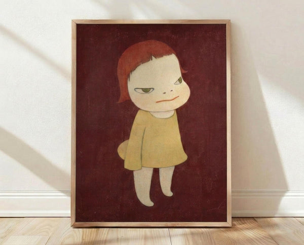 May include: Framed art print displaying a stylized illustration of a pale child-like figure with reddish-orange hair and green eyes, wearing a yellow dress against a deep burgundy background. The artwork is shown in a light wood frame, leaning against a white wall on wooden flooring with soft natural lighting creating shadows.