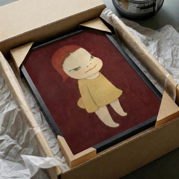 A framed poster featuring a minimalist illustration of a young child with red-orange hair wearing a yellow dress against a deep burgundy background, shown unpacked in a cardboard shipping box with brown paper wrapping and wooden frame corner protectors visible around the black frame.