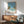 May include: A framed vintage-style poster showing a serene pool scene with palm trees and blue sky, displayed above a wooden mid-century modern credenza with sliding doors. A minimalist white ceramic table lamp sits on the credenza, and a gray upholstered armchair with wooden frame is positioned nearby. Warm natural light creates soft shadows on the white wall, giving the room a calm, retro aesthetic with teal and warm wood tones throughout.