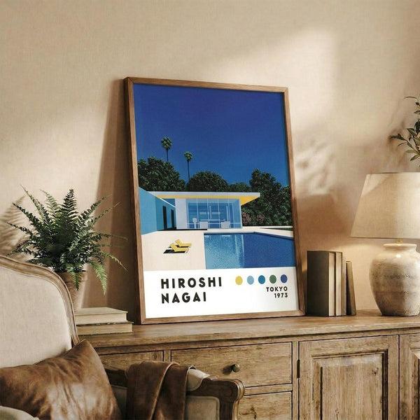 May include: Framed poster showing modernist house with blue pool against clear sky and palm trees, displayed on wooden sideboard with books, ceramic vase, and fern plant in cozy living room setting with beige armchair and neutral walls