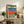 May include: Framed poster artwork displayed on rustic wooden console table in modern living room setting. The artwork shows a poolside scene with lounge chairs, blue water, orange diving board, and person wearing headphones sitting by rocky area. Natural wood frame, styled with fern plant, books, ceramic vase, and beige furniture visible in background.