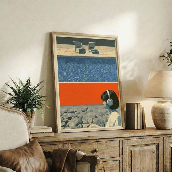 May include: Framed poster artwork displayed on rustic wooden console table in modern living room setting. The artwork shows a poolside scene with lounge chairs, blue water, orange diving board, and person wearing headphones sitting by rocky area. Natural wood frame, styled with fern plant, books, ceramic vase, and beige furniture visible in background.