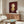 May include: Framed poster of a stylized child character with red-orange hair and yellow dress on deep burgundy background, displayed above a mid-century modern wooden credenza with sliding doors. Room features white curved chair, oval glass coffee table, white mushroom lamp, and brass floor mirror against cream walls with natural lighting.