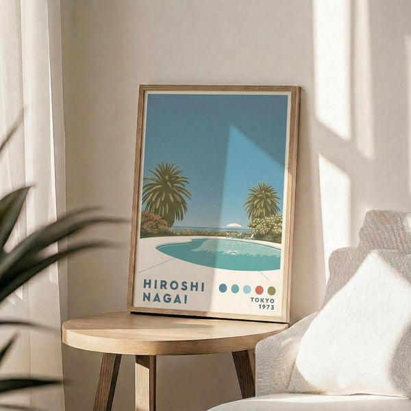 May include: Framed art print displayed on round wooden side table next to beige upholstered chair. Print shows poolside scene with turquoise swimming pool, two palm trees, and blue sky in retro illustration style. Text reads "Hiroshi Nagai" and "Tokyo 1973" with colorful dots. Natural wood frame, bright sunlight streaming across white wall, green plant leaves visible in corner.