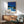 May include: Framed poster displaying modern architectural illustration of minimalist house with swimming pool, palm trees, and bright blue sky, hanging above wooden sideboard with table lamp, next to mid-century modern green upholstered armchair in contemporary living room setting with natural lighting casting shadows on white wall.